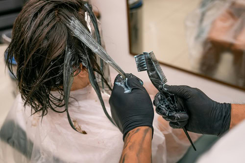 A person with shoulder-length dark hair sits in a salon chair, covered by a white cape. A stylist wearing black gloves applies a keratin treatment with a brush, mindful of potential hair salon chemical exposure during the keratin smoothing process. Read more at Aerovex Systems (aerovexsystems.com)