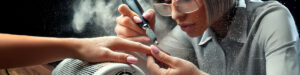 A nail technician wearing glasses shapes a clients nails with an electric nail file, using a nail dust collector that is releasing harmful nail dust into the air, illustrating the question, do cheap nail dust collectors work. The client’s nails are painted light pink. The scene is close-up and focused on the hands and tools used during the manicure. Read more at Aerovex Systems (aerovexsystems.com)