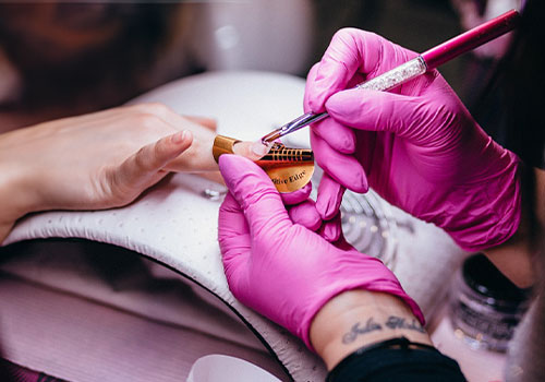 A person wearing pink gloves applies gel or acrylic to another person's fingernail using a brush. The hand receiving the manicure rests on a white cushion, and a nail extension form is placed under the fingernail. Nail products and tools are visible nearby. The focus is on the hand and nail work which indicates airborne VOCs for a discussion on the demand for cleaner nail salon air, with blurred background details. Read more at Aerovex Systems (aerovexsystems.com)