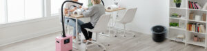 Two people sit across from each other at a manicure table in a bright, modern room. A pink Aerovex Whisper source capture air filtration machine with hose is beside them, ensuring room air purification along with the black The One That Works air purifier in the background. This illustrates the post question - Why combine source capture with room air purification? Read more at Aerovex Systems (aerovexsystems.com)