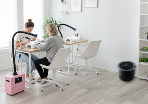 Two women sit at a manicure table in a bright, modern nail salon. One gives the other a manicure, with a pink Aerovex Whisper source capture air purification machine nearby and plants and wall art with a bookshelf and a The One That Works air purifier in the background. The room has light wood floors and white furniture. This illustrates the post question - Why combine source capture with room air purification? Read more at Aerovex Systems (aerovexsystems.com)