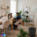 Three people work at a wooden table in a bright, modern nail studio filled with green plants. Soft pink chairs surround the table. Framed art decorates the white walls. A The One That Works air purifier sits on the light wood floor in the foreground. A gray Aerovex Systems Whisper Source Capture Systems sits between the two tables. It has two black flexible hoses with LED lit ends. Read more at Aerovex Systems (aerovexsystems.com)