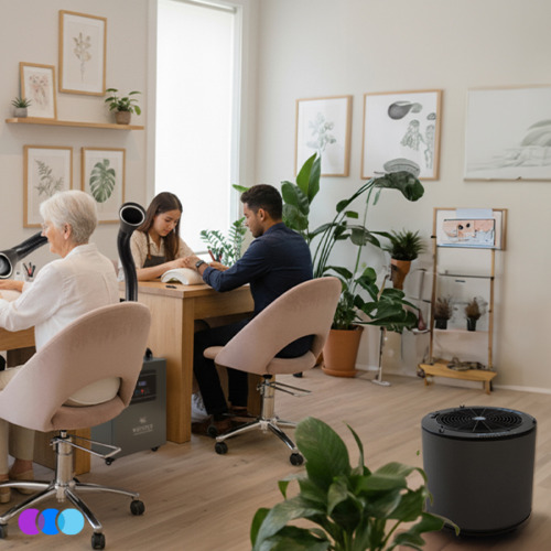 aerovex-whisper-dual-gray-the-one-that-works-totw-studio Three people work at a wooden table in a bright, modern nail studio filled with green plants. Soft pink chairs surround the table. Framed art decorates the white walls. A The One That Works air purifier sits on the light wood floor in the foreground. A gray Aerovex Systems Whisper Source Capture Systems sits between the two tables. It has two black flexible hoses with LED lit ends. Read more at Aerovex Systems (aerovexsystems.com)