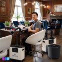 A man sits smiling at a nail salon workstation, surrounded by two Aerovex Systems Whisper Source Capture Systems, one with a two hose configuration and one with a single hose, LED light configuration. A The One That Works air purifier sits next to him on the floor. The elegant salon features wooden walls, large windows, paintings, and stylish lighting. Nail polish bottles are visible on the desk. Read more at Aerovex Systems (aerovexsystems.com)