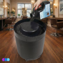 A hand pours black Aerovex Systems Activated Carbon Pellets into a The One That Works™ Salon Room Air Purifier by Aerovex Systems on a wooden floor visible near salon chairs and mirrors in the softly blurred background. Read more at Aerovex Systems (aerovexsystems.com)