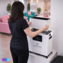 A woman with shoulder-length brown hair, wearing a black t-shirt and jeans, places a new filter set into a white Aerovex Whisper™ salon source capture system with a digital display. The setting is a modern, brightly lit room with pink chairs and decorative shelves. Read more at Aerovex Systems (aerovexsystems.com)