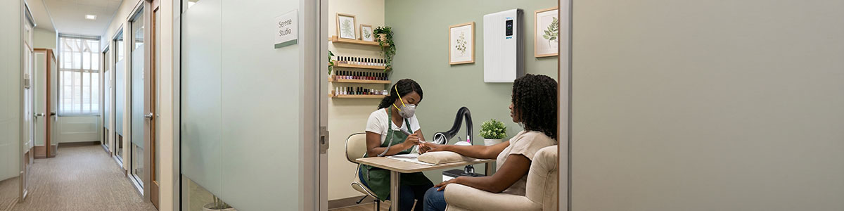 A nail technician wearing an Airwave 4800 N95 mask works on a customer's nails in a well-lit, modern nail studio. Shelves with nail polish and framed art decorate the space. Both women are seated at a manicure table in a clean, organized room. A white Aerovex MiniVent single station with LED light is actively suctioning debris and fumes from the breathing zone while an Aerovex Max wall mounted air purifier cleans the room air. Read more at Aerovex Systems (aerovexsystems.com)
