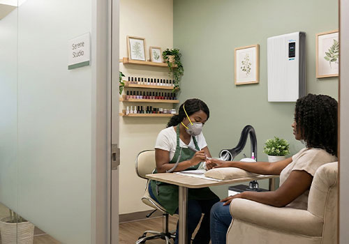 A nail technician wearing an Airwave 4800 N95 mask works on a customer's nails in a well-lit, modern nail studio. Shelves with nail polish and framed art decorate the space. Both women are seated at a manicure table in a clean, organized room. A white Aerovex MiniVent single station with LED light is actively suctioning debris and fumes from the breathing zone while an Aerovex Max wall mounted air purifier cleans the room air. Read more at Aerovex Systems (aerovexsystems.com)