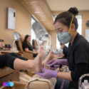 A nail technician wearing a mask and gloves gives a pedicure to a client in a salon. A fume extractor hovers near the client’s feet, capturing dust. Other clients and technicians are visible in the background, all seated in a well-lit, clean setting. Read more at Aerovex Systems (aerovexsystems.com)