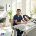 Wearing an Airwave 4800 N95 Mask Plus Relief From Organic Vapors, navy scrubs, and pink gloves, a person sits at a manicure station in a bright nail salon with an Aerovex Whisper dust collector in pink for organic vapors, plants, desk lamp, and sunlight from large windows. An Aerovex Max Wall Mounted Air Purifier is seen on the back wall with shelves of nail polish. Read more at Aerovex Systems (aerovexsystems.com)
