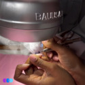 A close-up of hands performing a manicure. One hand holds an electric nail file shaping acrylic nails on the other hand, under a large silver nail dust collector labeled "BAUUSA." The workspace has a pink surface and scattered manicure tools. Read more at Aerovex Systems (aerovexsystems.com)