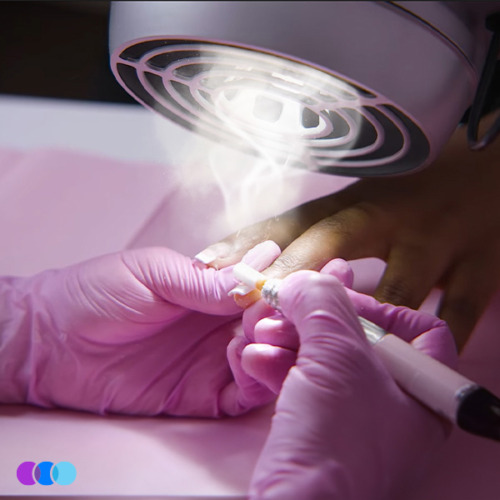 aerovex-sunflower-ii-manicure-action-3 A close-up of gloved hands performing a manicure with an electric nail file under a vented lamp. White dust rises as the tool shapes a fingernail. The background is pink and purple, with a small logo of three overlapping circles in the corner. Read more at Aerovex Systems (aerovexsystems.com)