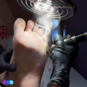 A gloved hand uses an electric tool to file the bottom of a person’s foot under a vent with visible suction, removing dead skin. The foot is held steady, and the setting appears clinical. Three colored circles are seen at the bottom left corner. Read more at Aerovex Systems (aerovexsystems.com)