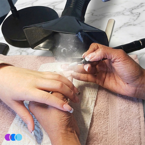 aerovex-whisper-single-action A nail technician uses an electric nail file on a client's fingernails, producing white dust. Both hands rest on a towel over a marble surface, with a ventilation device above to capture the dust. Nail products and tools are visible nearby. Read more at Aerovex Systems (aerovexsystems.com)