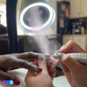 A close-up of a person using an electric nail file on another person’s fingernail in a well-lit room. Nail dust is being suctioned through the air into the Aerovex Whisper MiniVent™ source capture system with LED light. Both people have neatly manicured nails with different colored polish. Read more at Aerovex Systems (aerovexsystems.com)