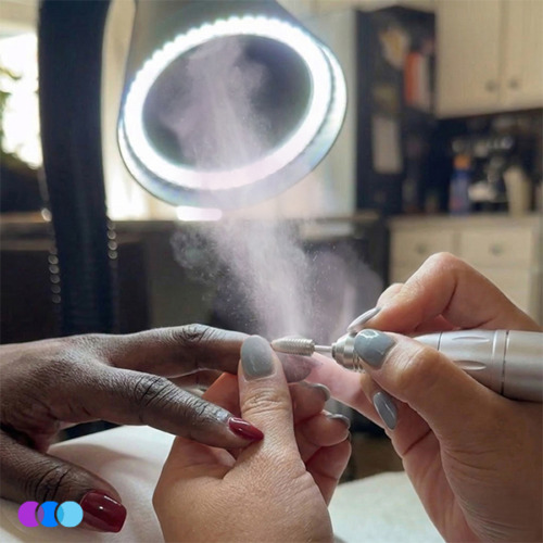 aerovex-whisper-single-led-action A close-up of a person using an electric nail file on another person’s fingernail in a well-lit room. Nail dust is being suctioned through the air into the Aerovex Whisper MiniVent™ source capture system with LED light. Both people have neatly manicured nails with different colored polish. Read more at Aerovex Systems (aerovexsystems.com)