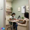 A nail technician wearing a mask files the nails of a client seated across a small table in a clean, well-lit nail salon. The salon has pastel green walls, framed art, shelves with nail polish, and a white air purifier on the wall. Read more at Aerovex Systems (aerovexsystems.com)