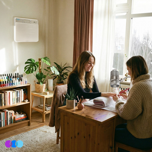 aerovex-wmap-white-sunflower-ii-cozy-home-studio Two women sit at a wooden table by a sunlit window in a cozy, plant-filled living room. One gives the other a manicure using an Aerovex Sunflower II Nail Dust Collector. A white Aerovex Wall Mounted Air Purifier is mounted on the wall above an end table with plants and a bookshelf with various nail polish bottles arranged neatly in rows on top. Read more at Aerovex Systems (aerovexsystems.com)