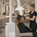 A hairstylist uses a straightener on a seated woman's hair in a modern salon. A large white air filtration device with a flexible hose hovers above the client, emitting gentle steam. Salon mirrors, chairs, and bright lighting are visible in the background. Read more at Aerovex Systems (aerovexsystems.com)