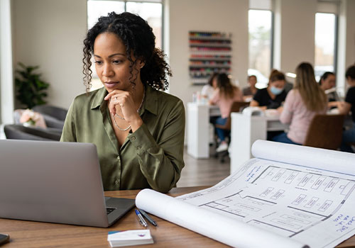 A woman with curly hair, wearing a green blouse, works on her laptop amid blueprints and business cards in a modern nail salon. Behind her, colleagues - some masked - discuss an IMC compliant nail salon source capture ventilation system while working with clients. Read more at Aerovex Systems (aerovexsystems.com)