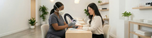 A nail technician wearing an Airwave N95 face mask and gloves gives a manicure to a seated woman in a modern, bright nail salon with plants, shelves of products, and white walls with an Aerovex Whisper MiniVent nail source capture system vaccuming the debris and VOCs away from their breathing zone. Both women are focused on the hands being worked on. Read more at Aerovex Systems (aerovexsystems.com)