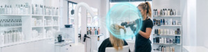 A hairstylist blow dries a clients long blonde hair for a keratin hair smoothing treatment in a bright, modern salon with shelves of hair products. A blue hologram shield highlights advanced salon ventilation for keratin treatments, adding a futuristic touch to the scene. Read more at Aerovex Systems (aerovexsystems.com)