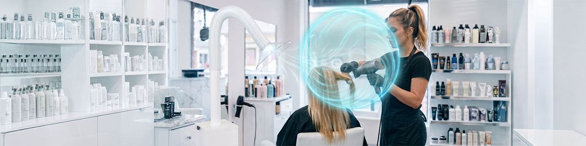 A hairstylist blow dries a clients long blonde hair for a keratin hair smoothing treatment in a bright, modern salon with shelves of hair products. A blue hologram shield highlights advanced salon ventilation for keratin treatments, adding a futuristic touch to the scene. Read more at Aerovex Systems (aerovexsystems.com)