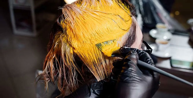 A person wearing black gloves applies bright yellow hair dye to someone’s hair with a brush—a scene familiar in hair and nail services, where source capture ventilation can help reduce inhalation risks from salon chemical fumes. Read more at Aerovex Systems (aerovexsystems.com)